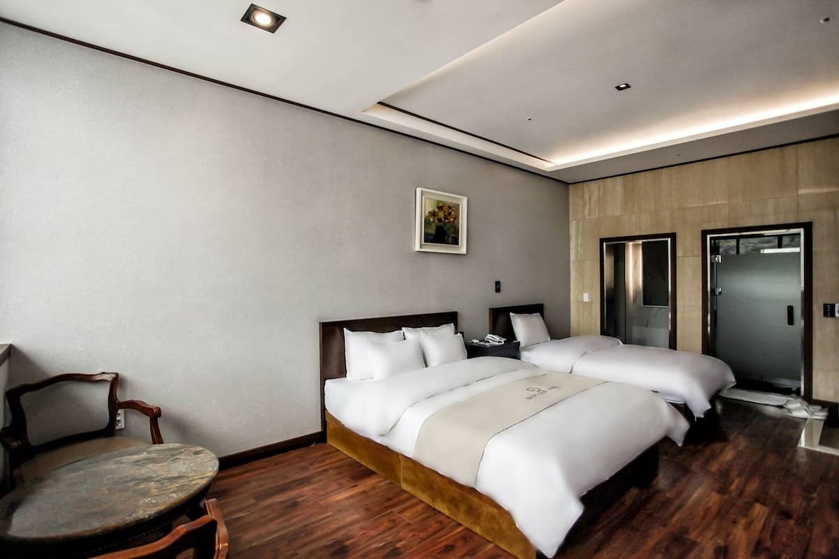 Habitación de hotel con piso de madera, dos camas con ropa blanca, una silla de madera, una mesita pequeña, un cuadro en la pared y una puerta con espejo.