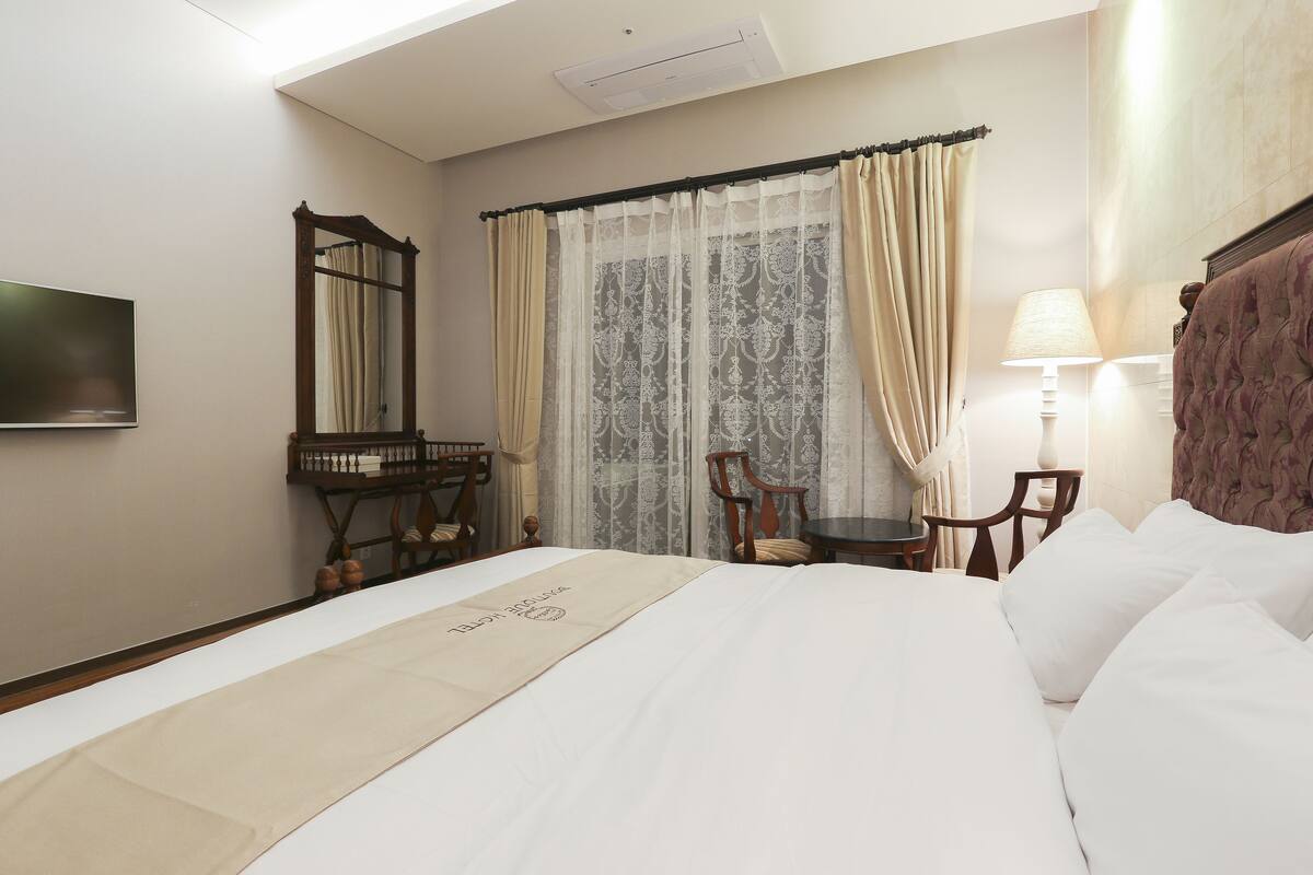 Habitación de hotel con cama, escritorio, silla, televisor y ventana con cortinas.