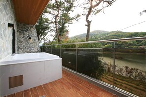 A balcony with a stone wall, a hot tub, and a view of a lake and hills. - Hotel Gallery (Pocheon)