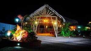 A thatched-roof structure with a wooden frame, surrounded by tropical plants and illuminated by ambient lighting.