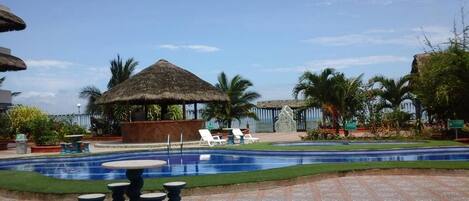 A pool area with lounge chairs, a thatched-roof structure, and palm trees.