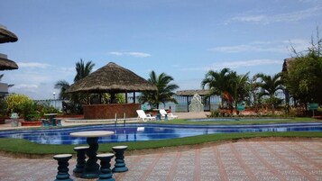 Un área de piscina con sillones reclinables, una estructura con techo de paja y palmeras.