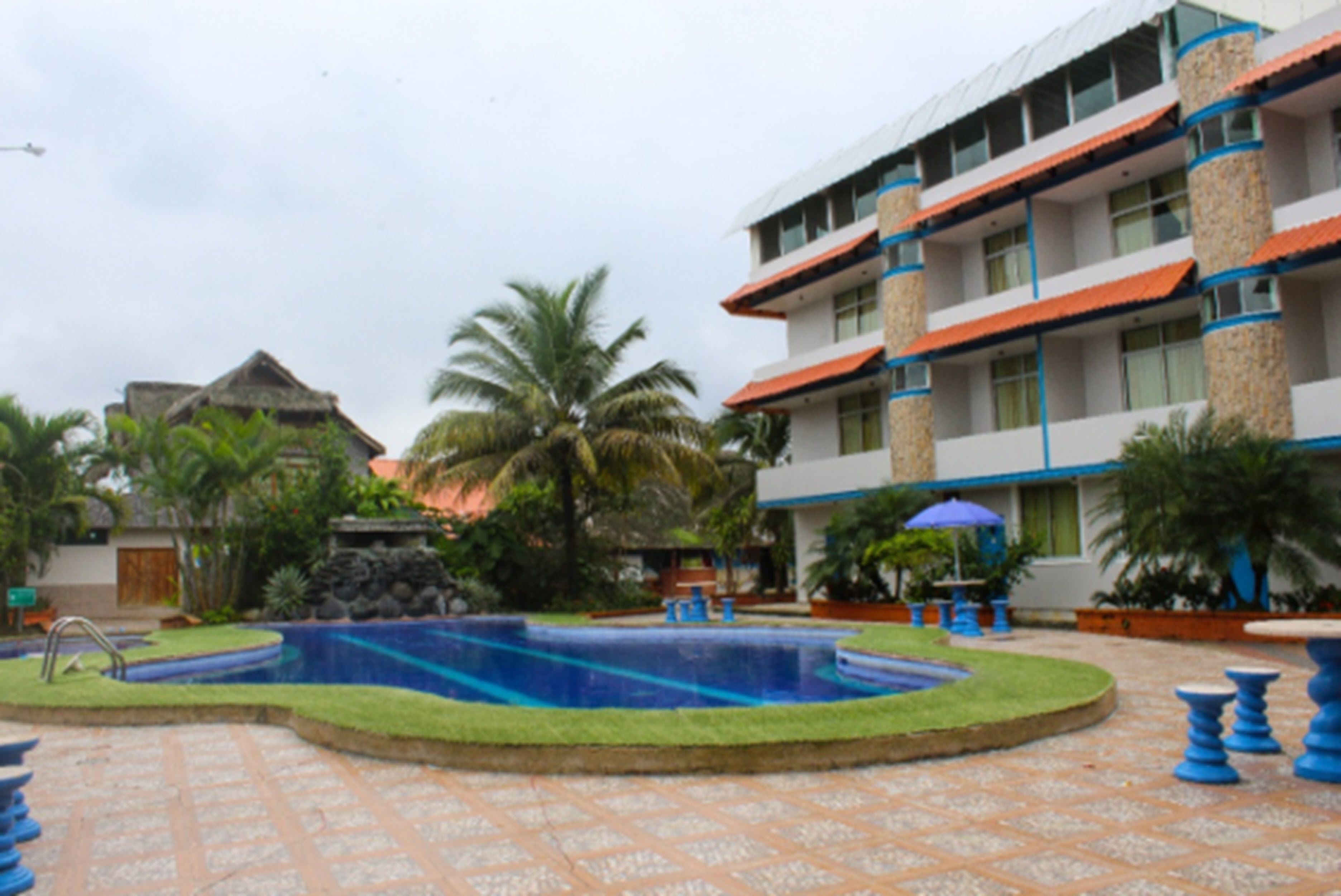 Una piscina rodeada por un jardín con palmeras, un edificio con varios balcones y un suelo de baldosas.