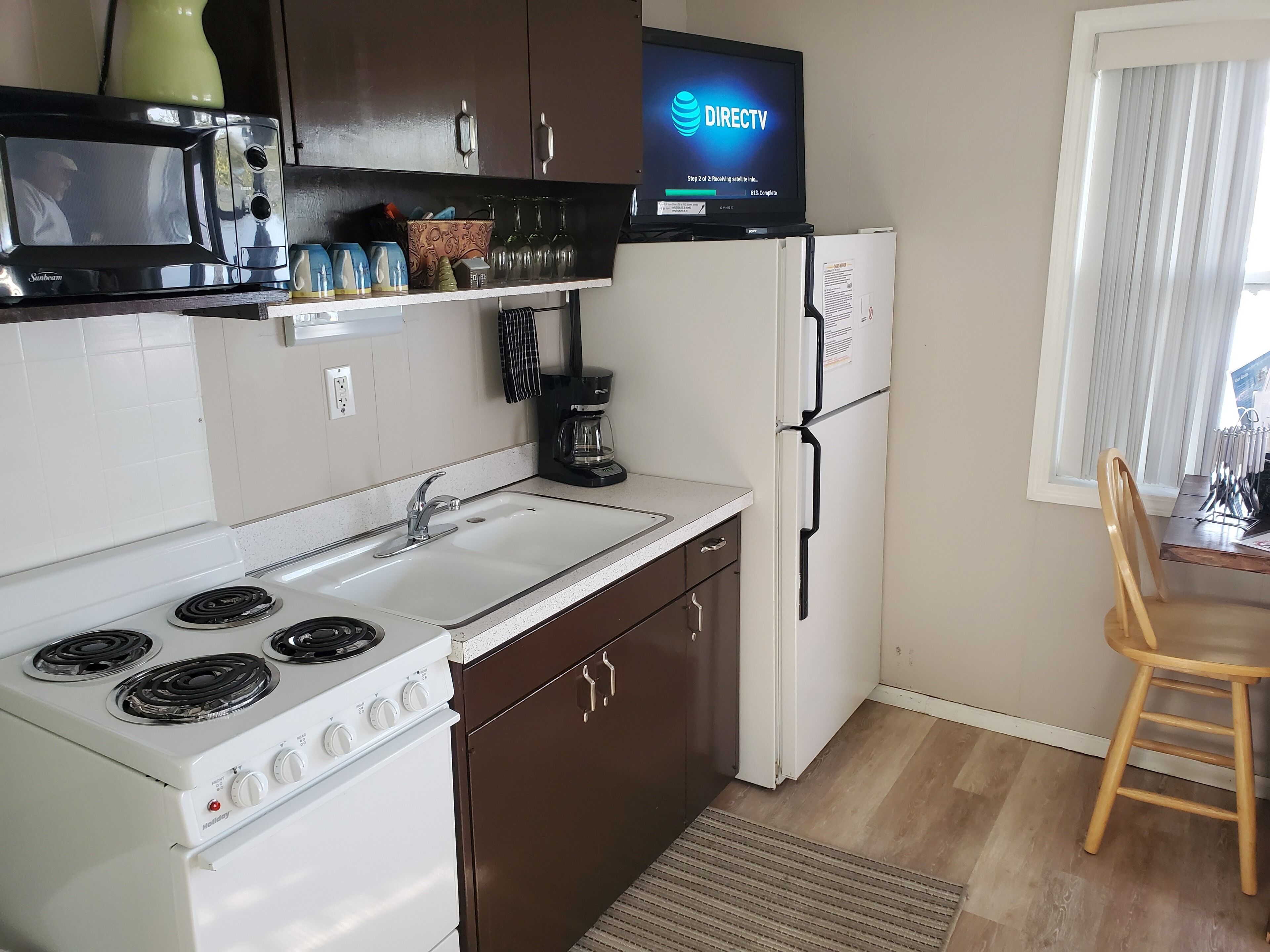 A compact kitchen with a stove, sink, and refrigerator, a flat-screen TV mounted above the counter, and a dining area with a chair.