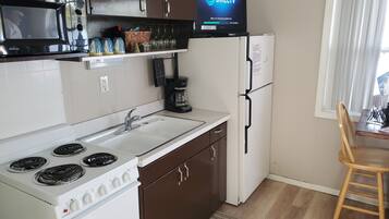 A compact kitchen with a stove, sink, and refrigerator, a flat-screen TV mounted above the counter, and a dining area with a chair.