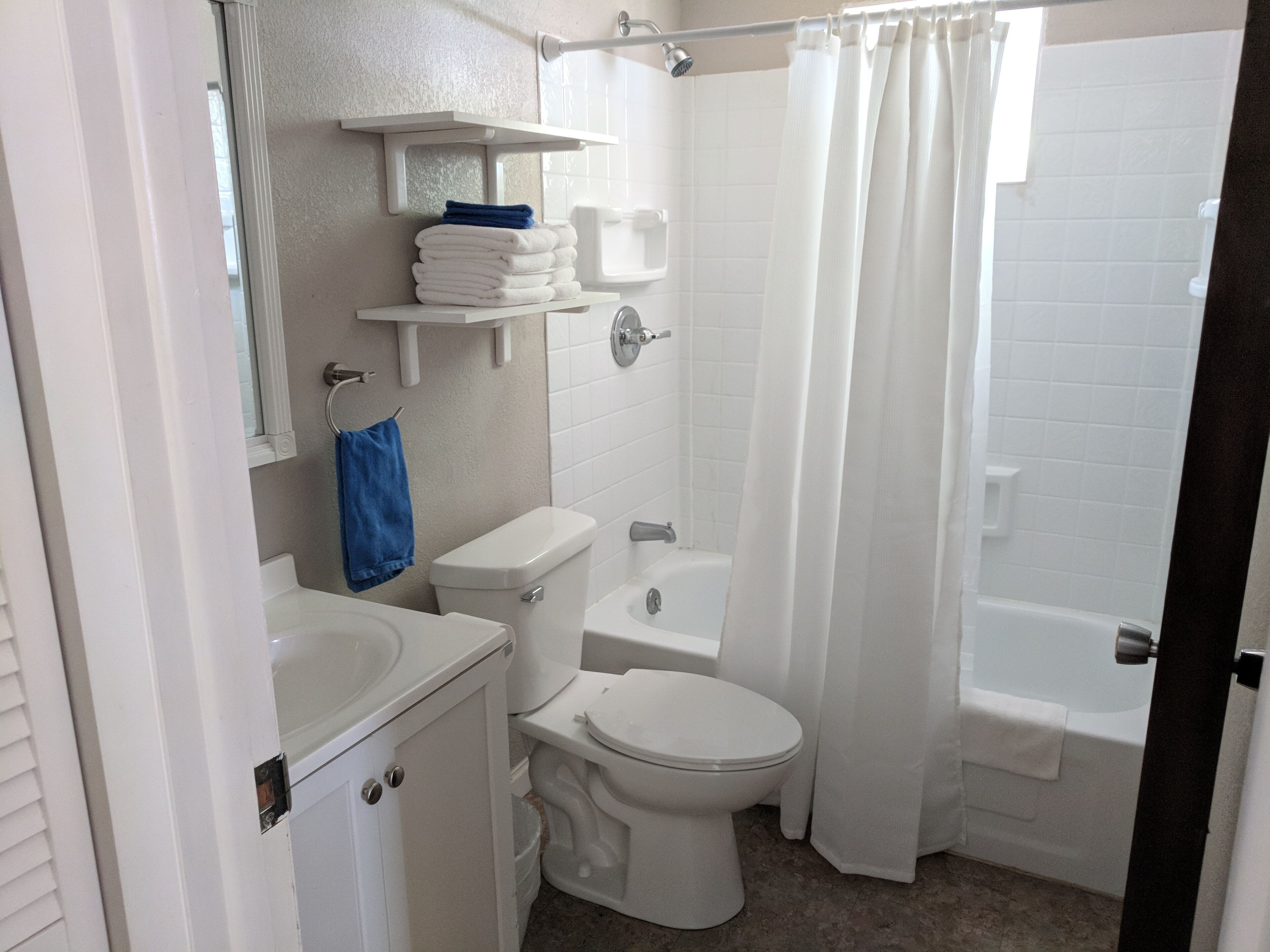 A bathroom with a toilet, sink, and bathtub. There are shelves above the toilet and a shower curtain.