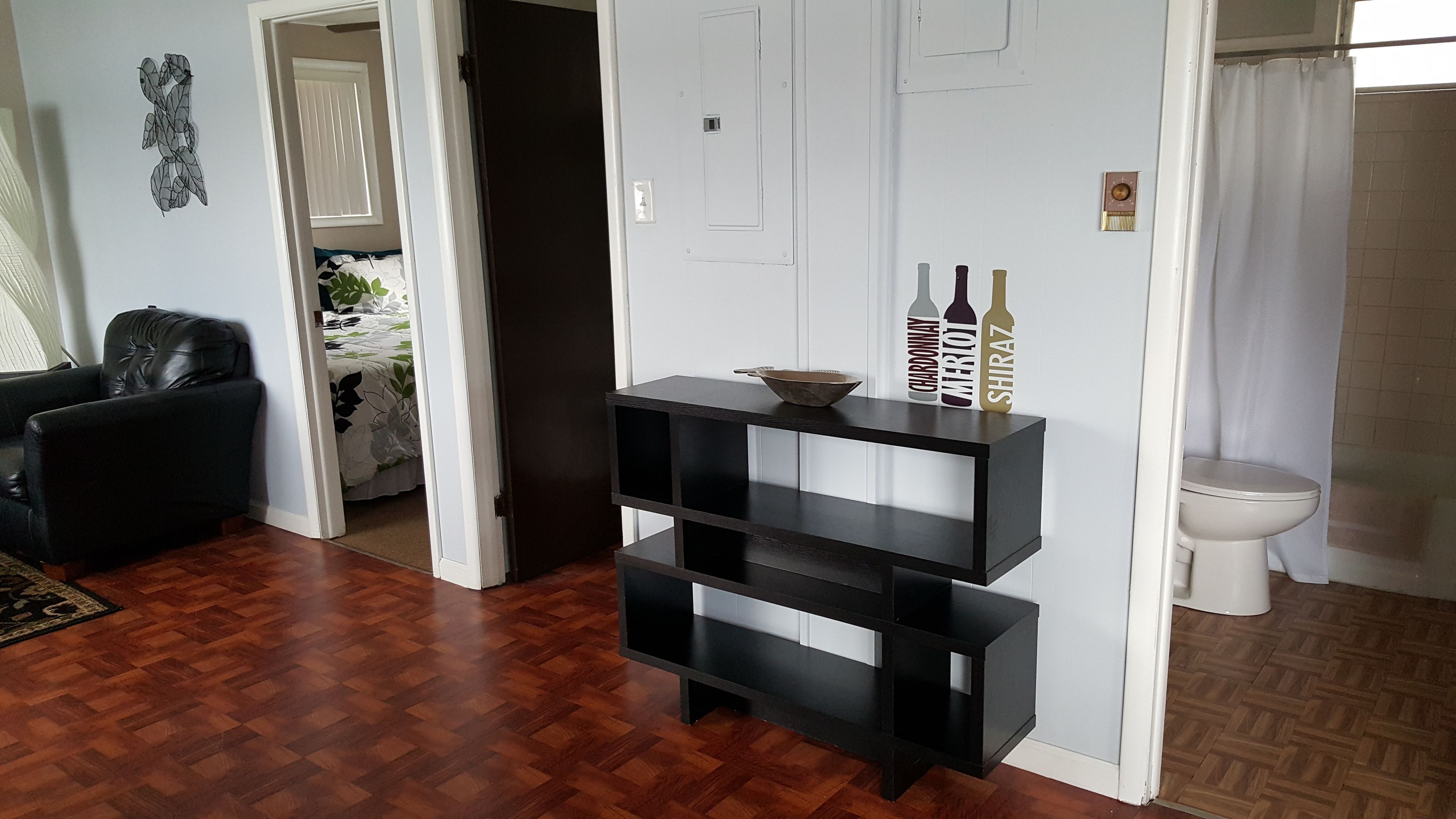 A living room with a dark wooden floor, a black leather chair, a white door, and a small black shelf with bottles.