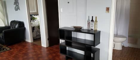 A living room with a dark wooden floor, a black leather chair, a white door, and a small black shelf with bottles.