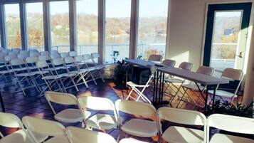 A conference room with white folding chairs arranged in rows, large windows with a view of trees, and a ceiling fan.