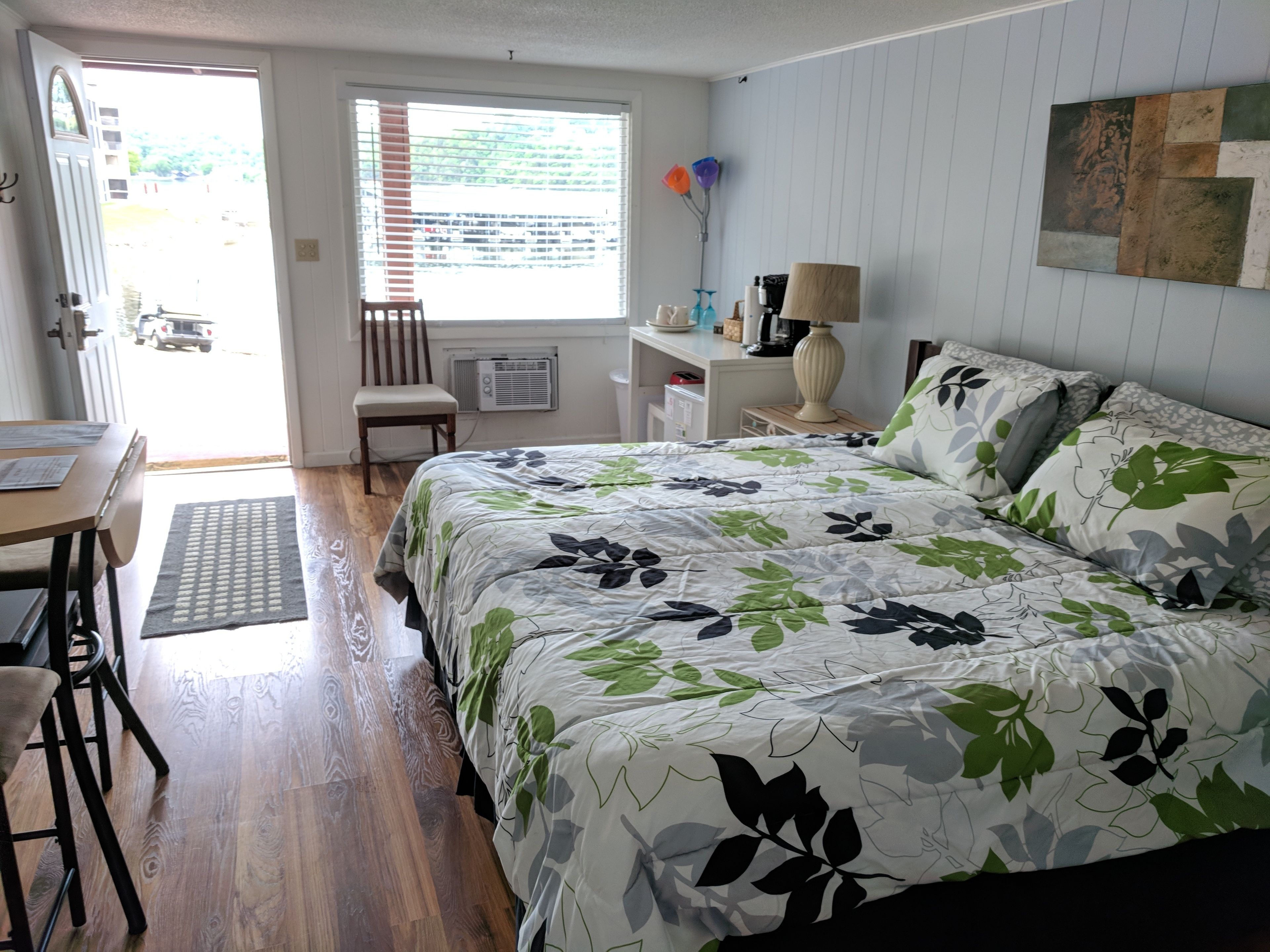 Une chambre avec un grand lit, un bureau et une chaise. Il y a une fenêtre avec des stores, un radiateur et une lampe sur une table de chevet.