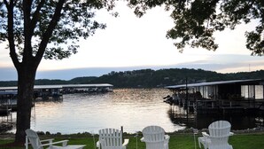 A lakeside view with docked boats, a tree, and white Adirondack chairs on the grass. - Lake Breeze Resort  (Camdenton)