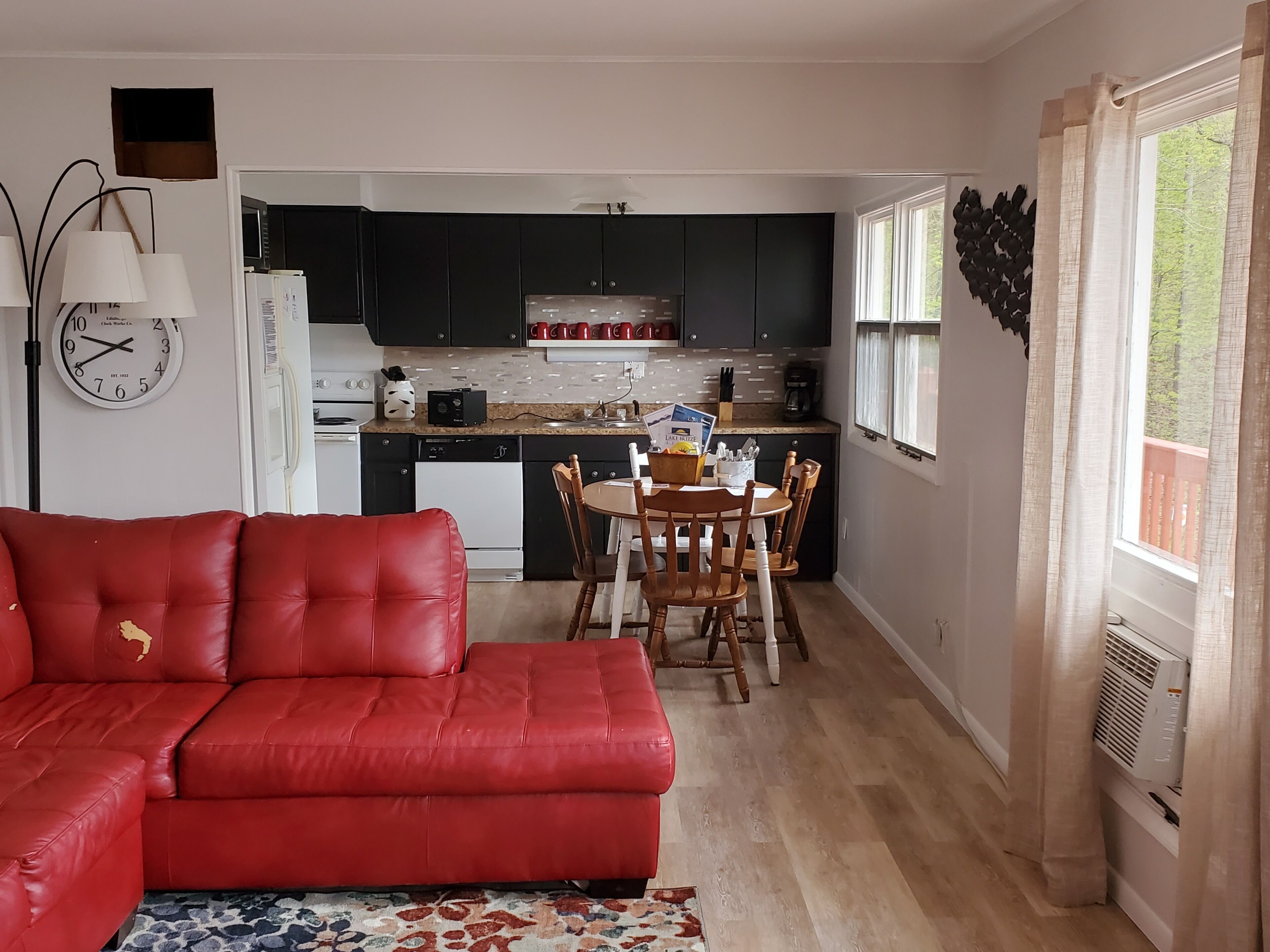 A compact living space with a red sofa, a dining table with chairs, a kitchen area with dark cabinets, a clock, and a window with curtains.