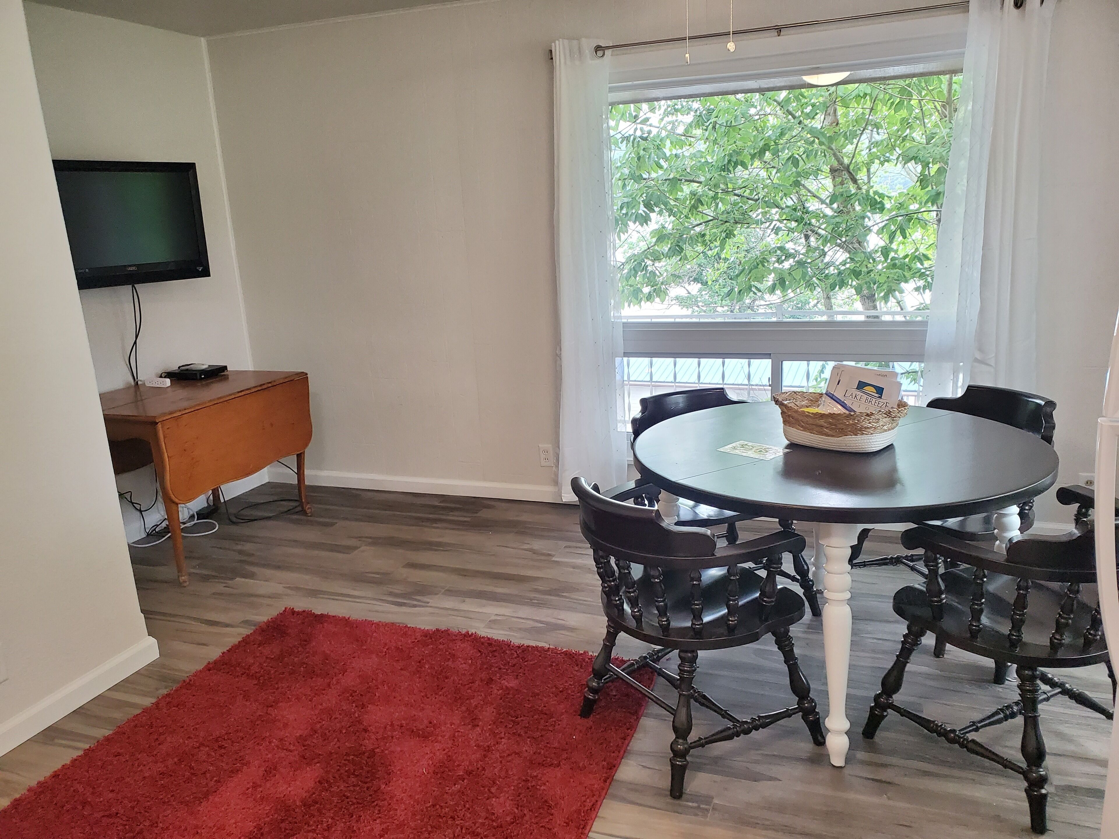 A dining area with a round table, black chairs, a red rug, a television on a stand, and a window with a view of trees.