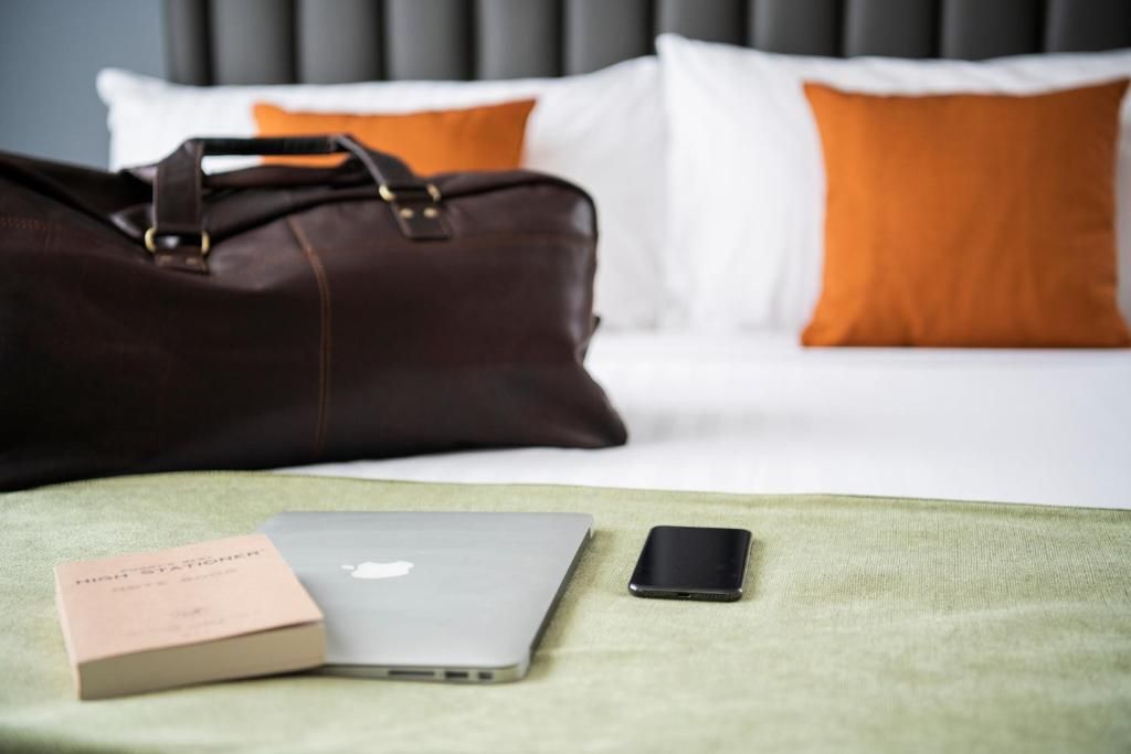 Un letto con una testiera grigia, cuscini bianchi e arancioni, una borsa in pelle marrone, un portatile, uno smartphone e un libro su una coperta verde.