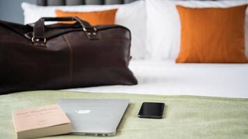 Un letto con una testiera grigia, cuscini bianchi e arancioni, una borsa in pelle marrone, un portatile, uno smartphone e un libro su una coperta verde.