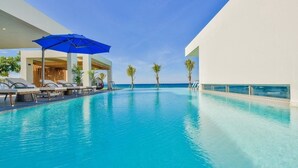 Une piscine entourée de chaises longues et de parasols, bordée de palmiers et surplombée d’un ciel dégagé.