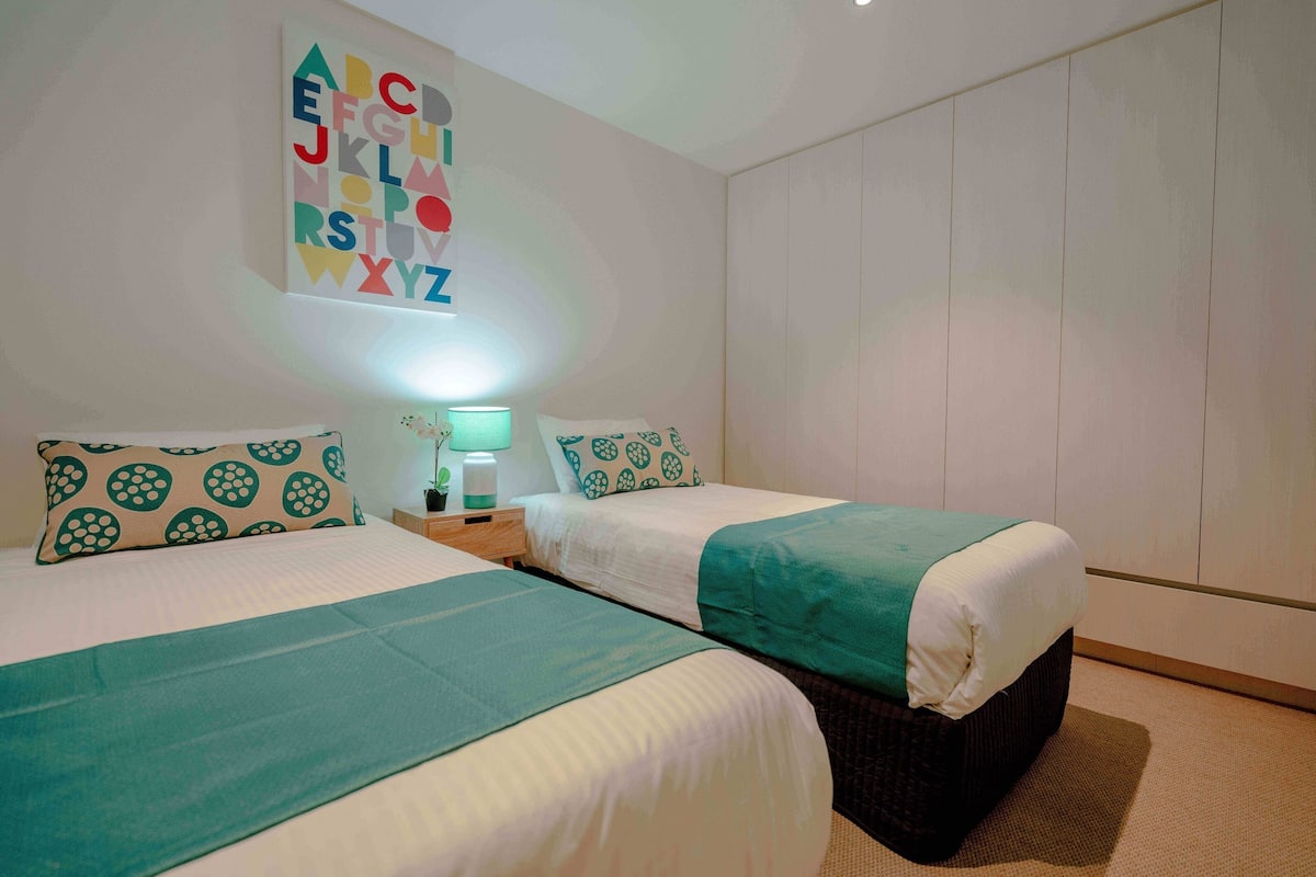 Habitación de hotel con dos camas, una pared con letras del alfabeto, una mesita de noche con lámpara y un ventanal grande.