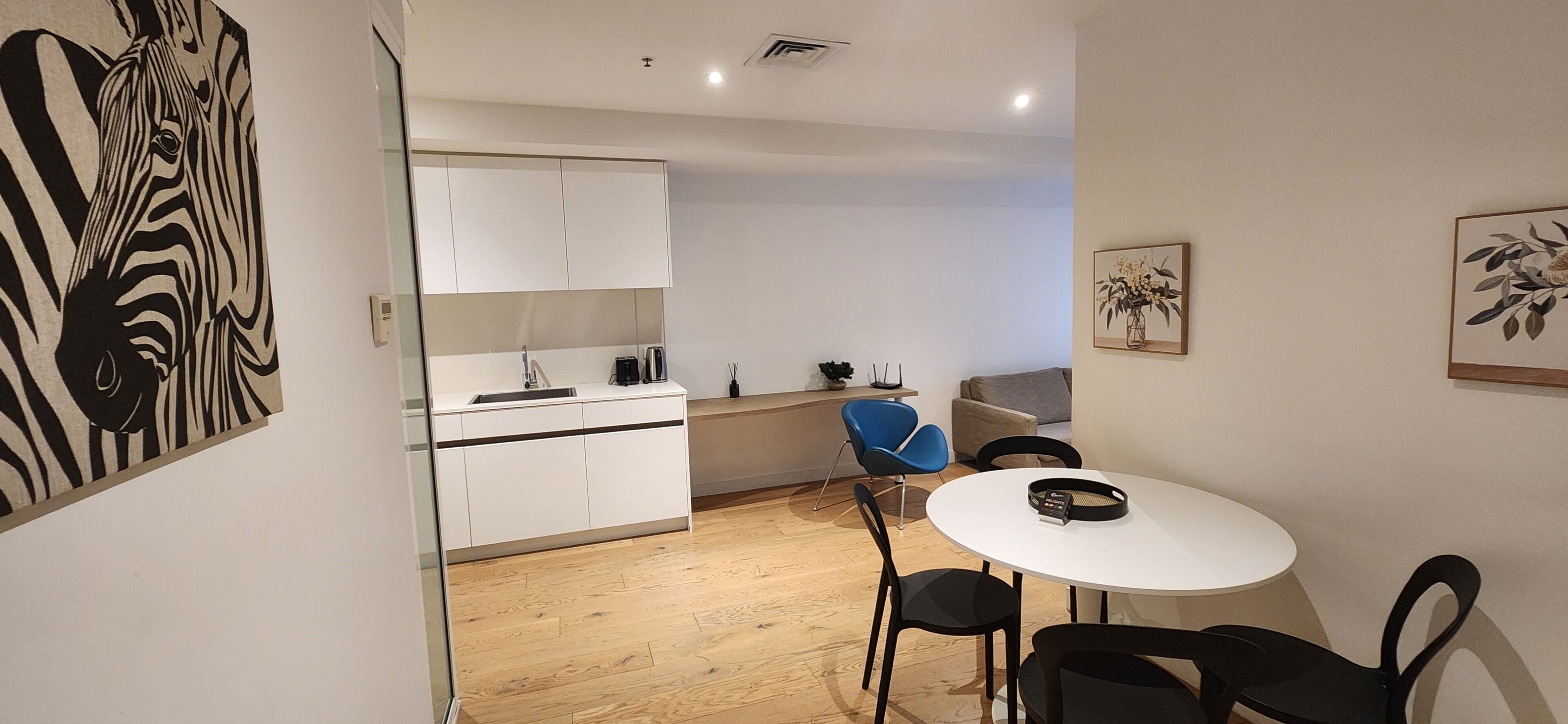A modern kitchen with white cabinets, a sink, and a coffee machine. There is a dining area with a white table and black chairs.