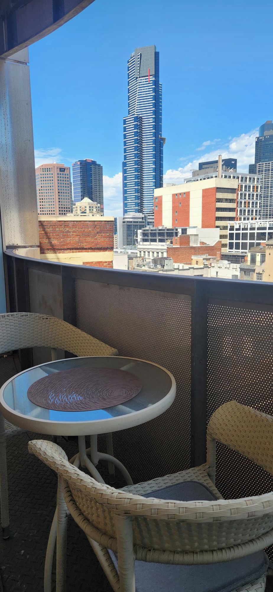 A balcony with wicker chairs and a small table, overlooking a cityscape with various buildings.
