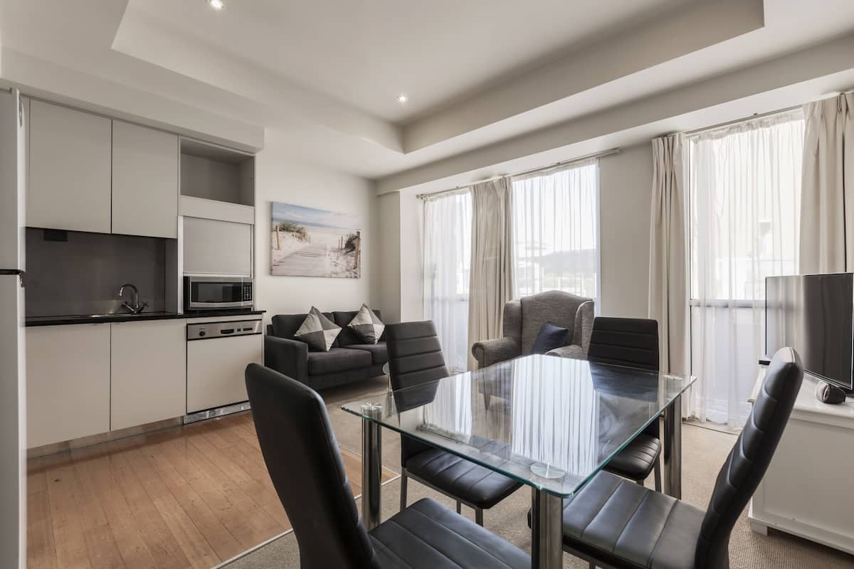 Una sala de estar moderna con una mesa de comedor de vidrio, sillas de cuero negro, un sofá gris y un televisor de pantalla plana sobre un mueble blanco.