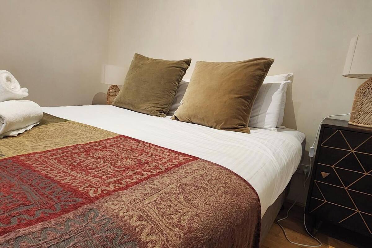 Una cama bien hecha con edredón blanco, dos almohadas marrones y un plaid rojo y beige.