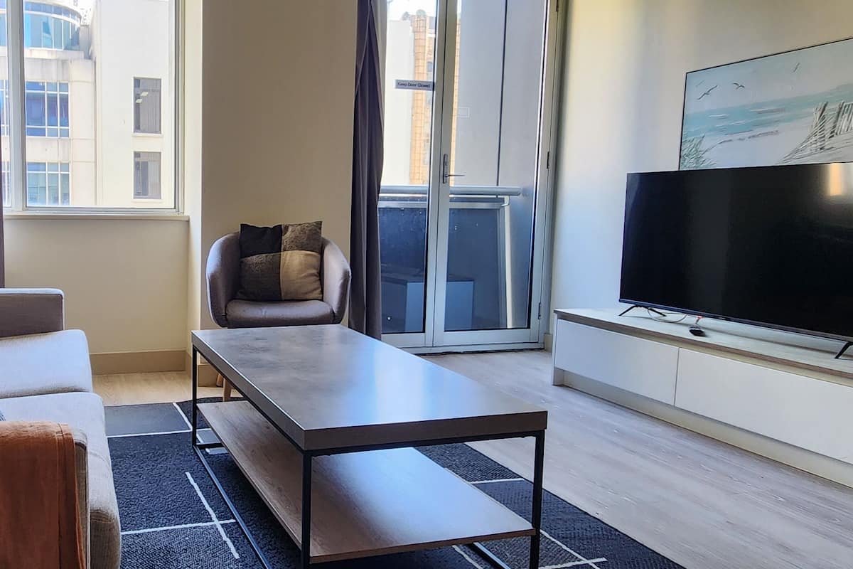 Una sala de estar moderna con un sofá, una mesa de centro, un televisor y vistas de edificios a través de una ventana.