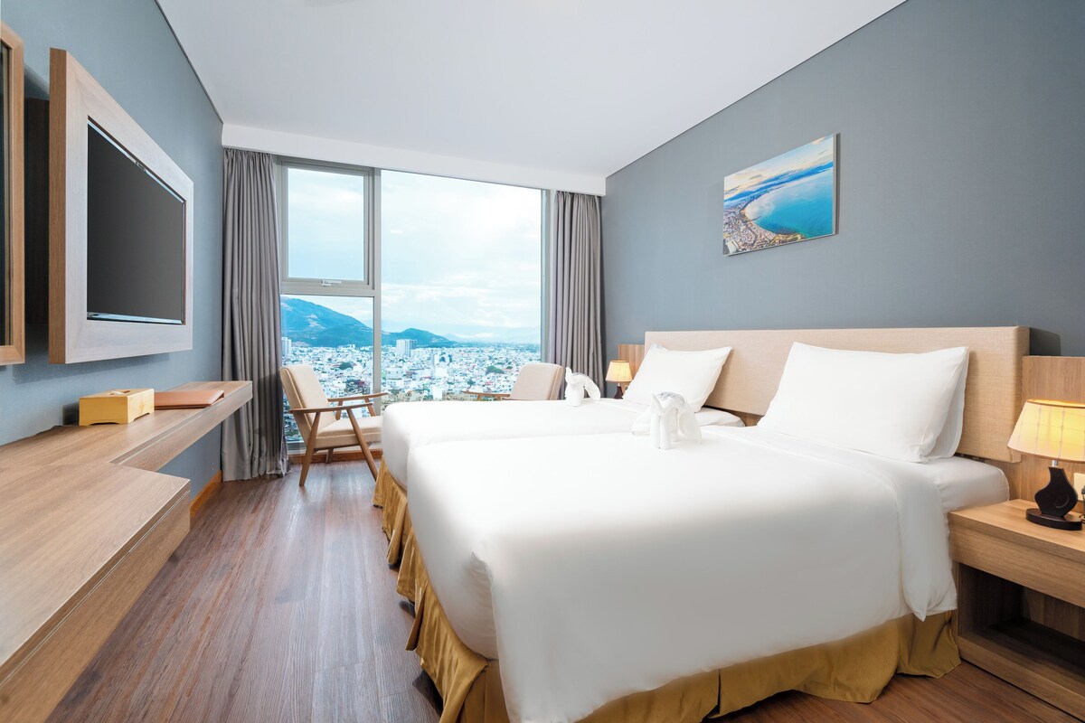 A hotel room with a large bed, a desk, a chair, a TV, and a window with a city and mountains view.