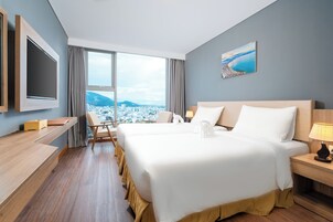 A hotel room with a large bed, a desk, a chair, a TV, and a window with a city and mountains view.
