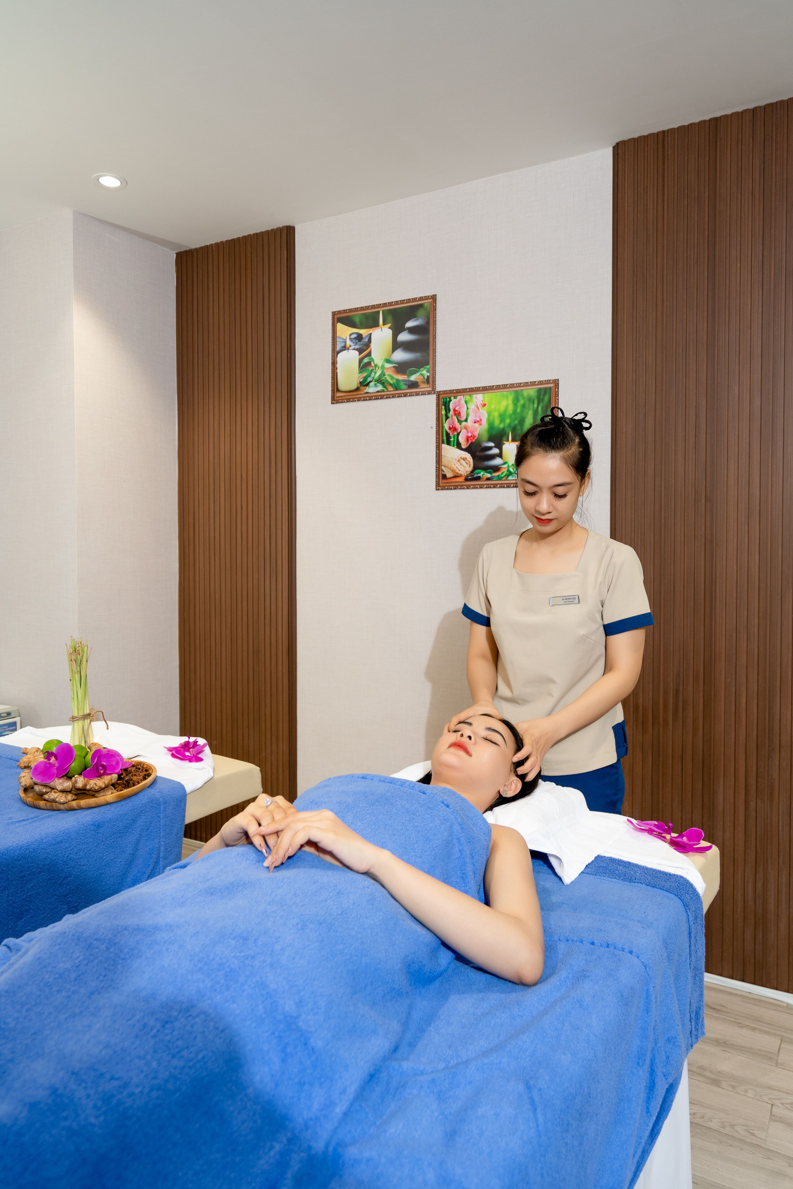 A massage therapy room with two massage tables, each covered with a blue towel. A massage therapist is attending to a client. The room has wooden paneling on the walls and a small table with a vase of flowers and a bowl of fruit.