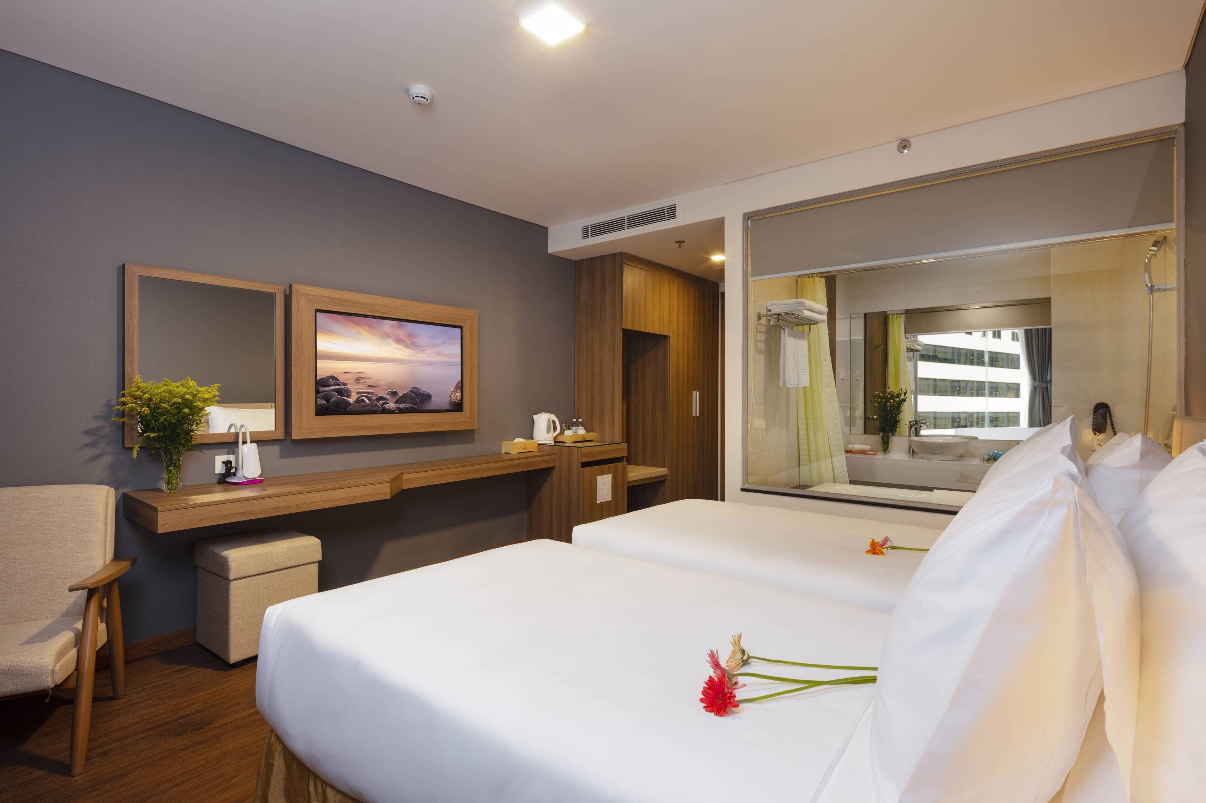 A hotel room with a bed, a desk with a computer, a chair, a small table with a vase of flowers, and a large mirror.