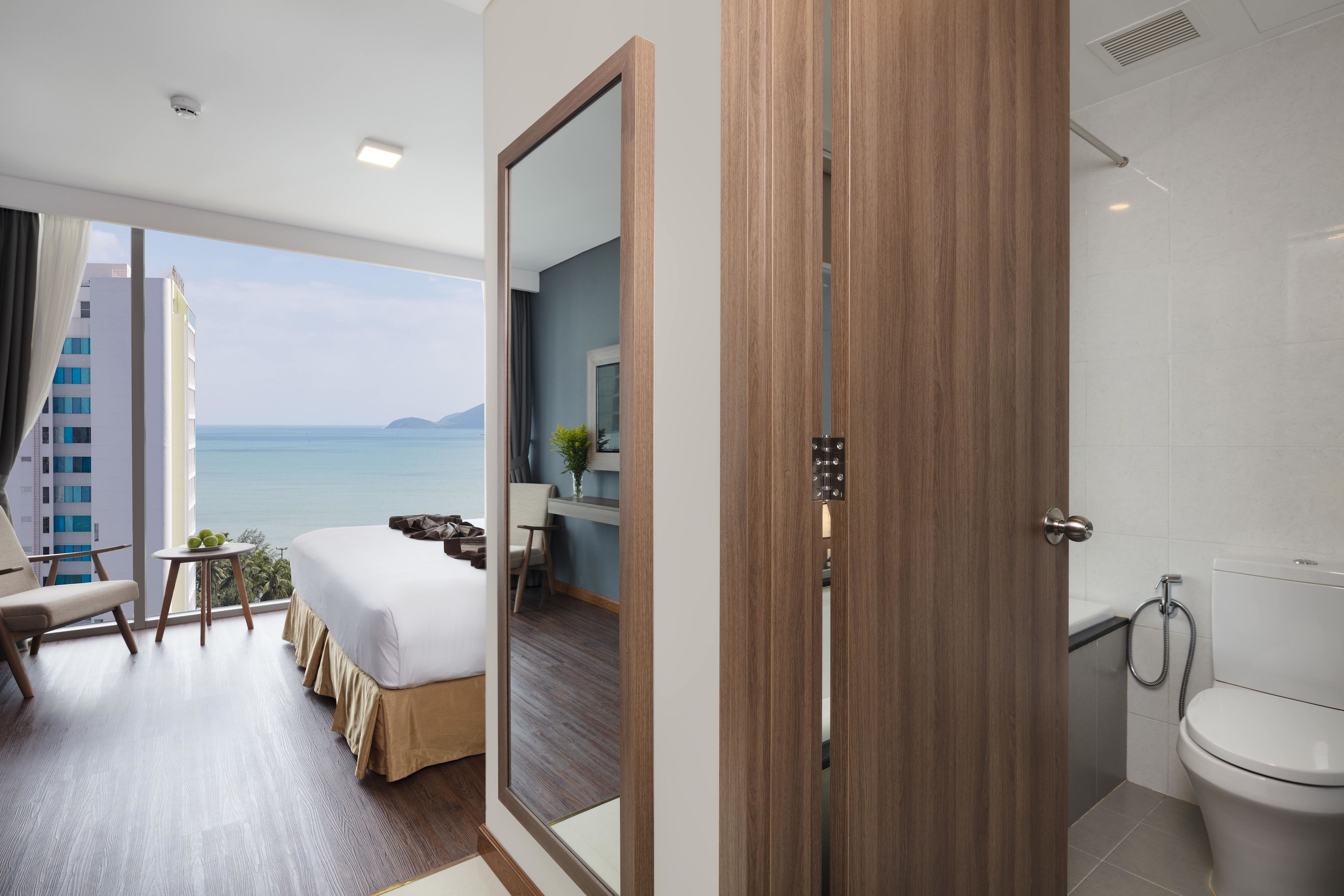 A hotel room with a bed, a seating area with a view of the sea, a bathroom with a toilet and shower, and a large mirror.