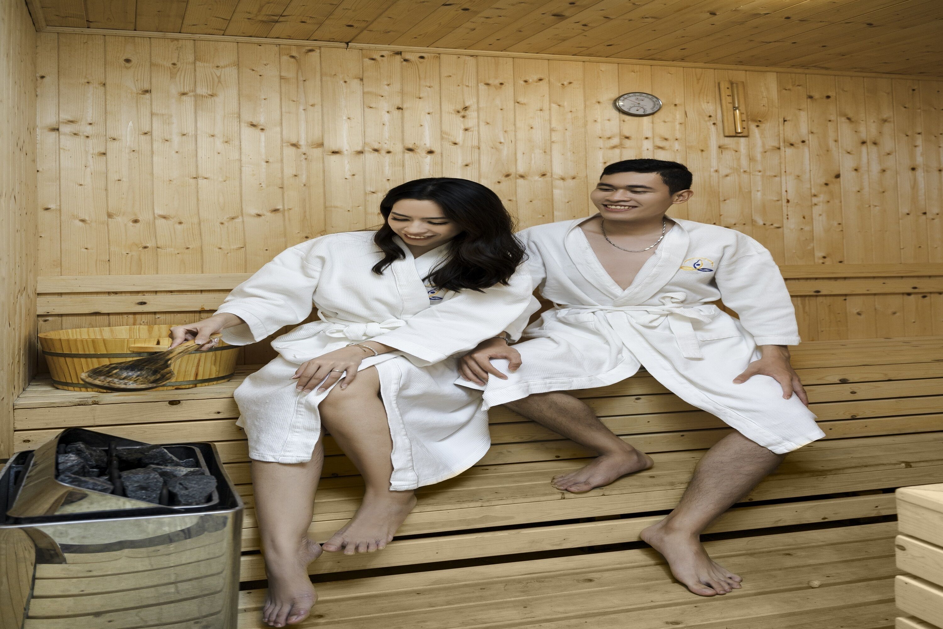 Zwei Personen in weißen Roben sitzen in einer Holzsauna mit einer hölzernen Sitzbank, einem Holzeimer und einem Mülleimer.