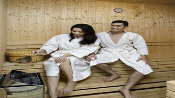 Zwei Personen in weißen Roben sitzen in einer Holzsauna mit einer hölzernen Sitzbank, einem Holzeimer und einem Mülleimer.