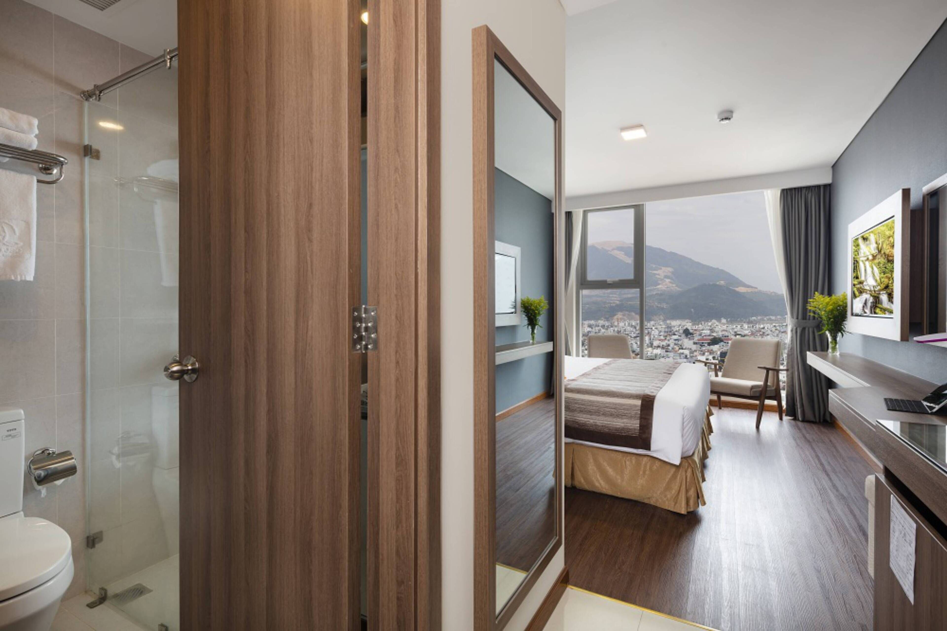 A modern hotel room with a large bed, a desk, and a view of a cityscape and mountains.