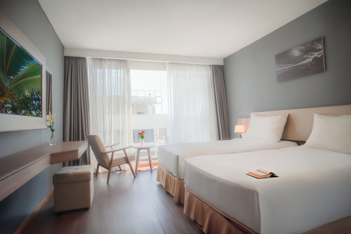 A hotel room with two beds, a desk, a chair, a small table with a vase, and a view of the city through large windows.