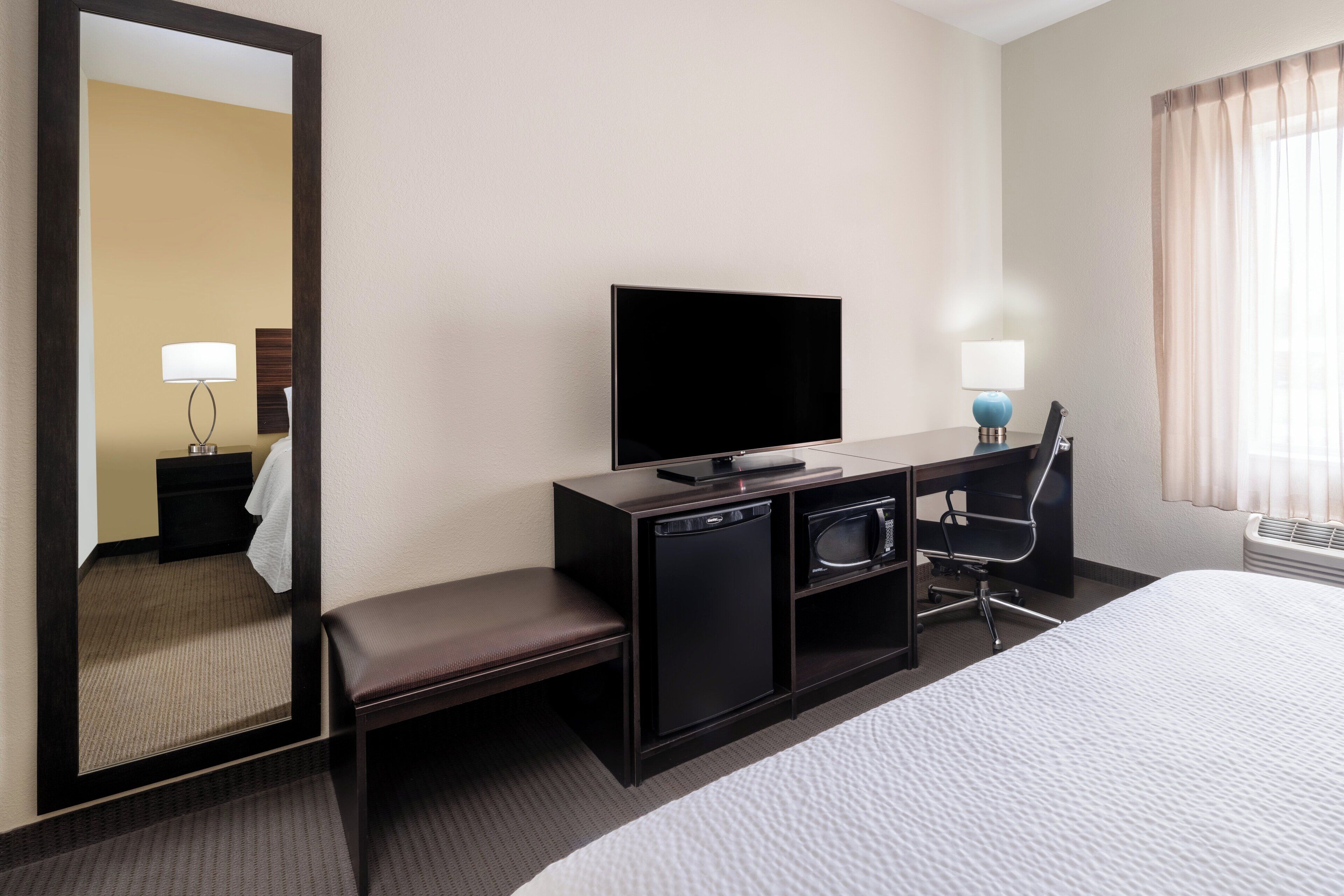 A hotel room with a bed, a desk with a flat-screen TV, a microwave, a chair, a bench, and a mirror.