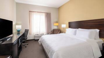 Habitación de hotel con cama, escritorio con televisor, silla, lámpara y ventana con cortinas.