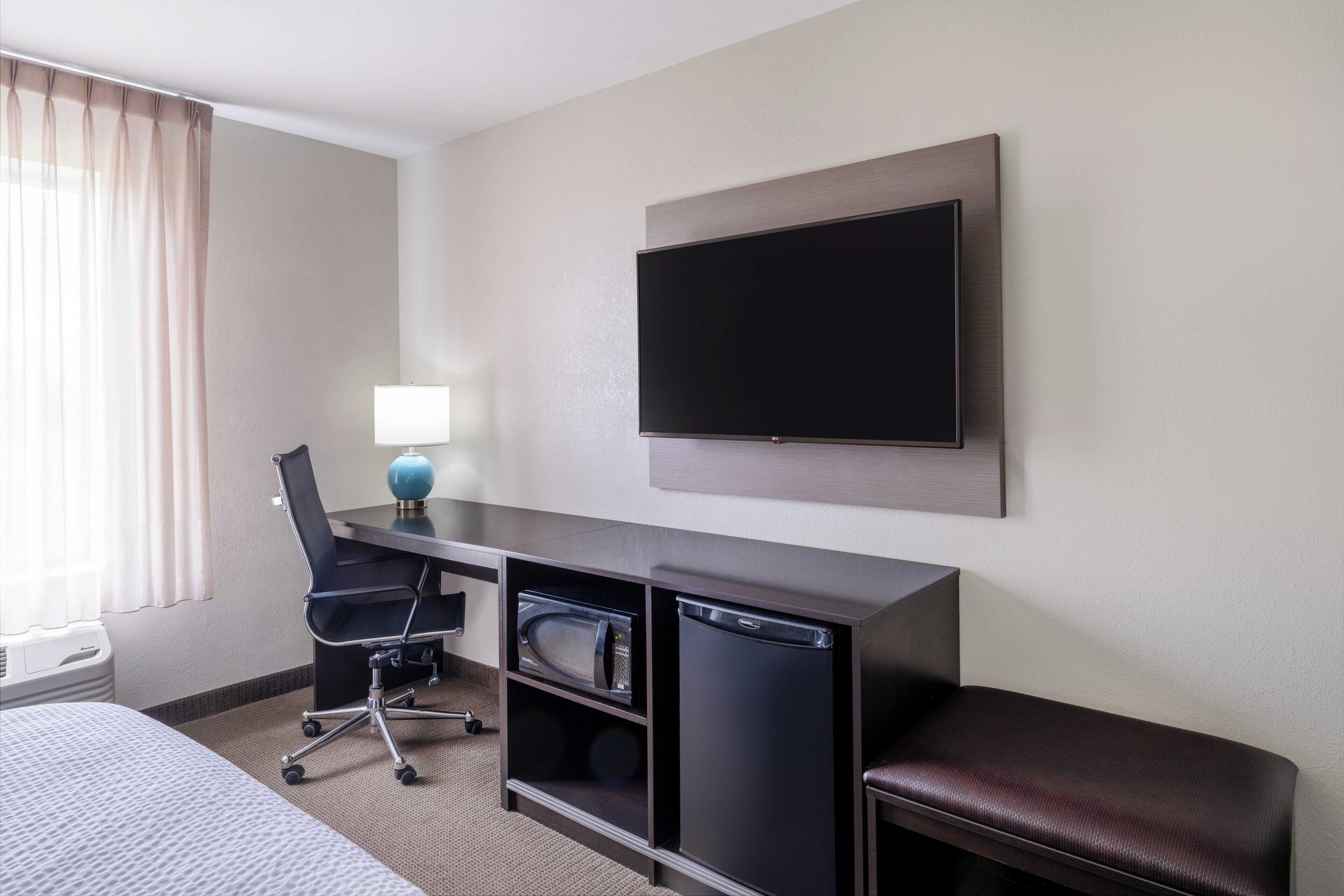 A hotel room with a bed, a desk with a computer, a microwave, a refrigerator, a TV, and a window with curtains.