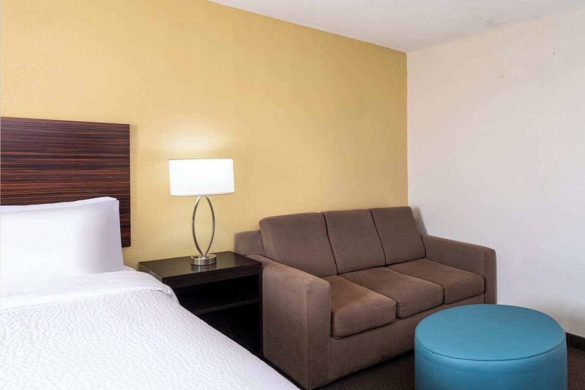 Una habitación de hotel con una cama, un sofá marrón, un reposapiés azul y una mesita de noche con lámpara.