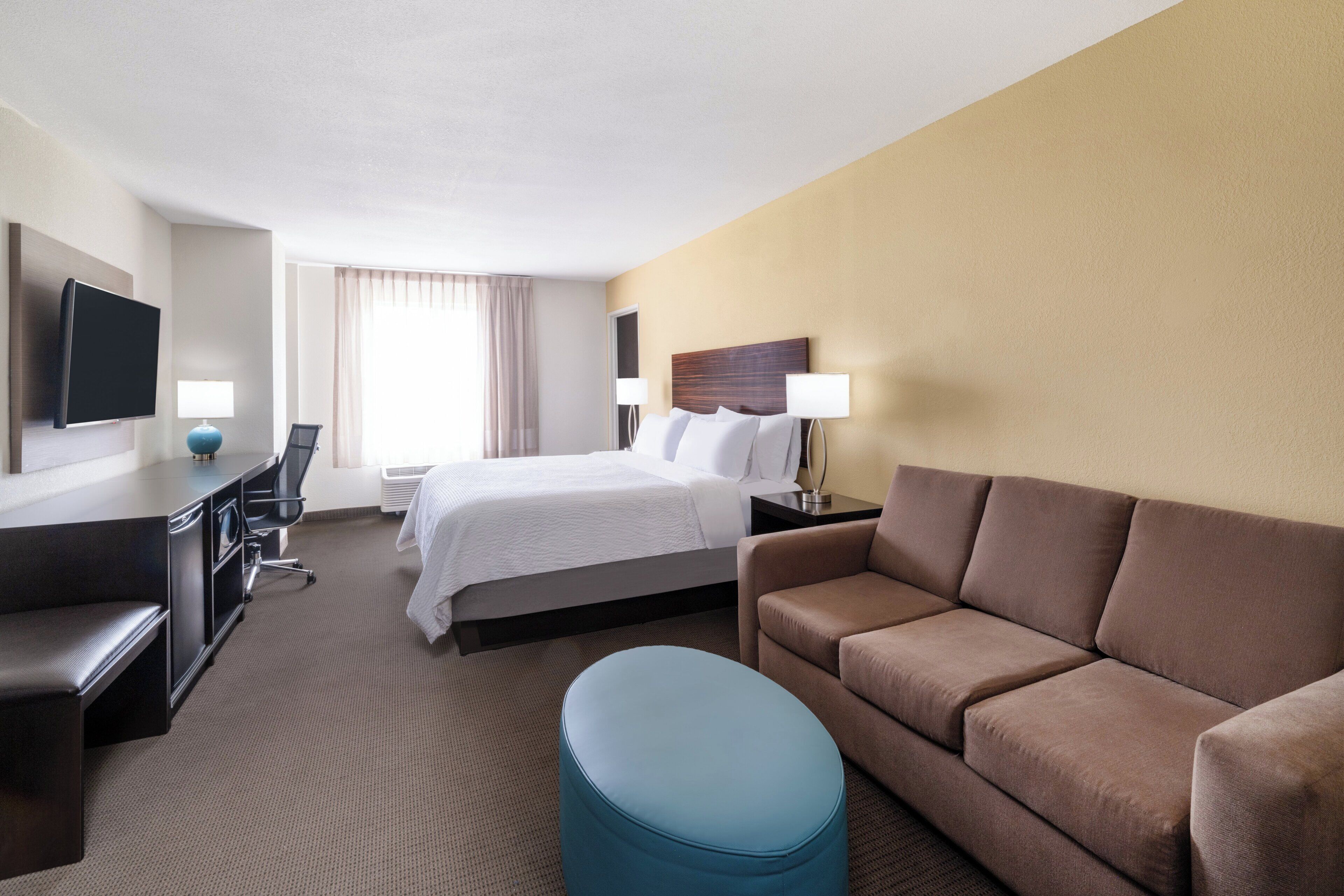 Une chambre d’hôtel avec un lit, un bureau, une télévision, un canapé et un pouf bleu.