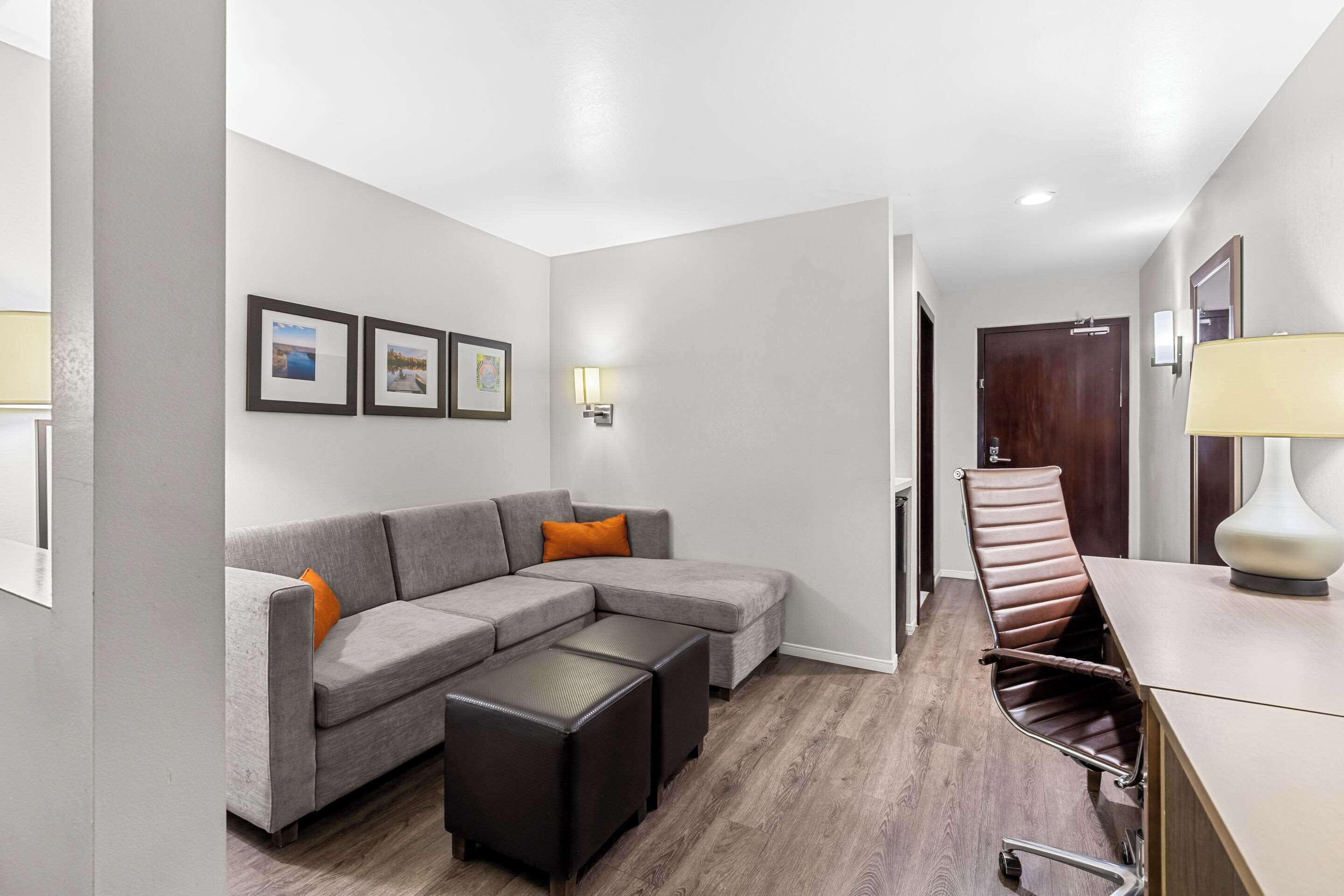 Een moderne woonkamer met een grijze hoekbank, een zwart poefje en een bruine leren fauteuil. Er hangen ingelijste foto's aan de muur en op de bijzettafel staat een lamp.