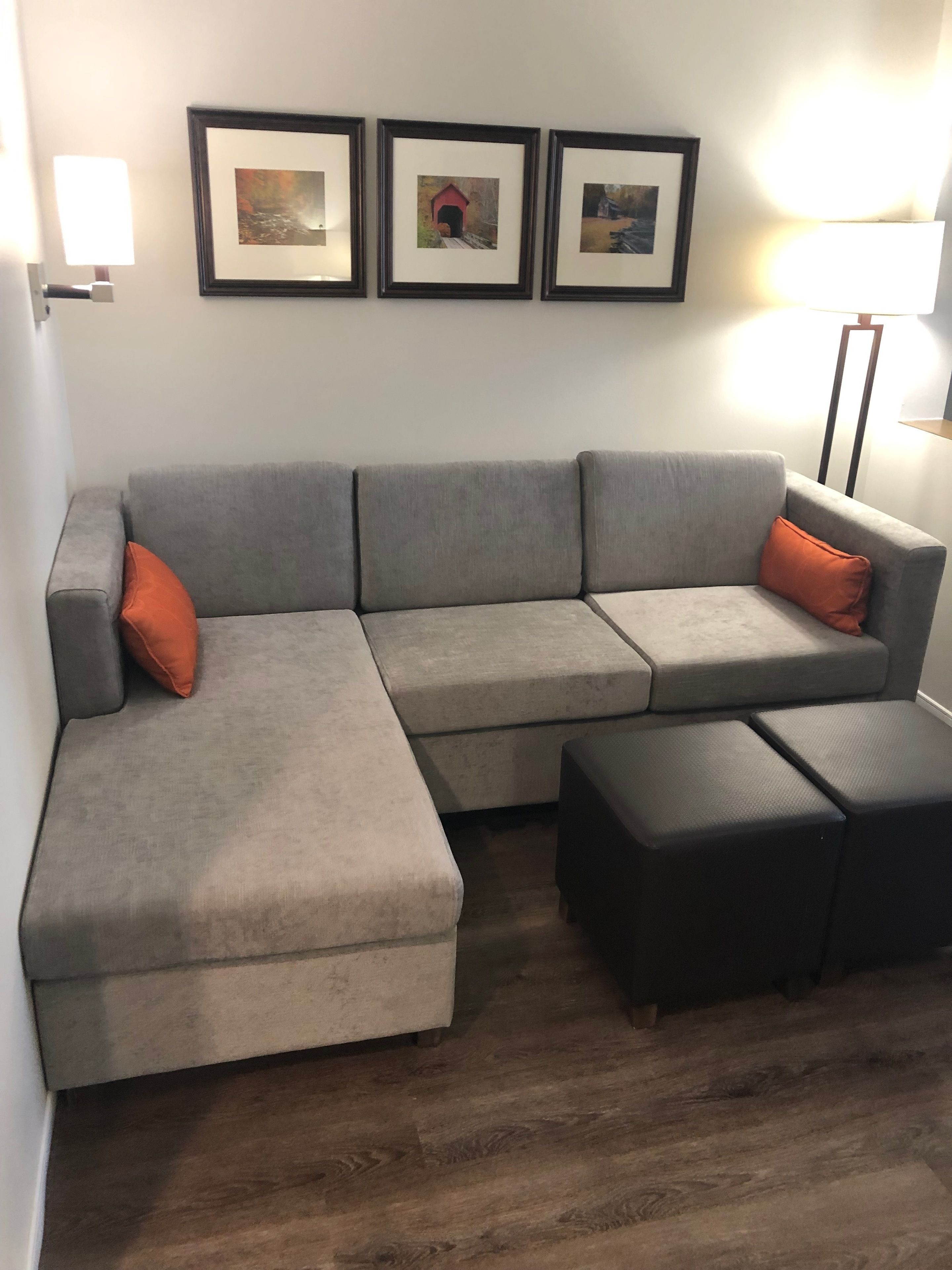 Ein graues Ecksofa mit orangefarbenen Kissen, ein schwarzer Hocker und drei gerahmte Bilder an der Wand.