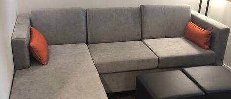 Ein graues Ecksofa mit orangefarbenen Kissen, ein schwarzer Hocker und drei gerahmte Bilder an der Wand.