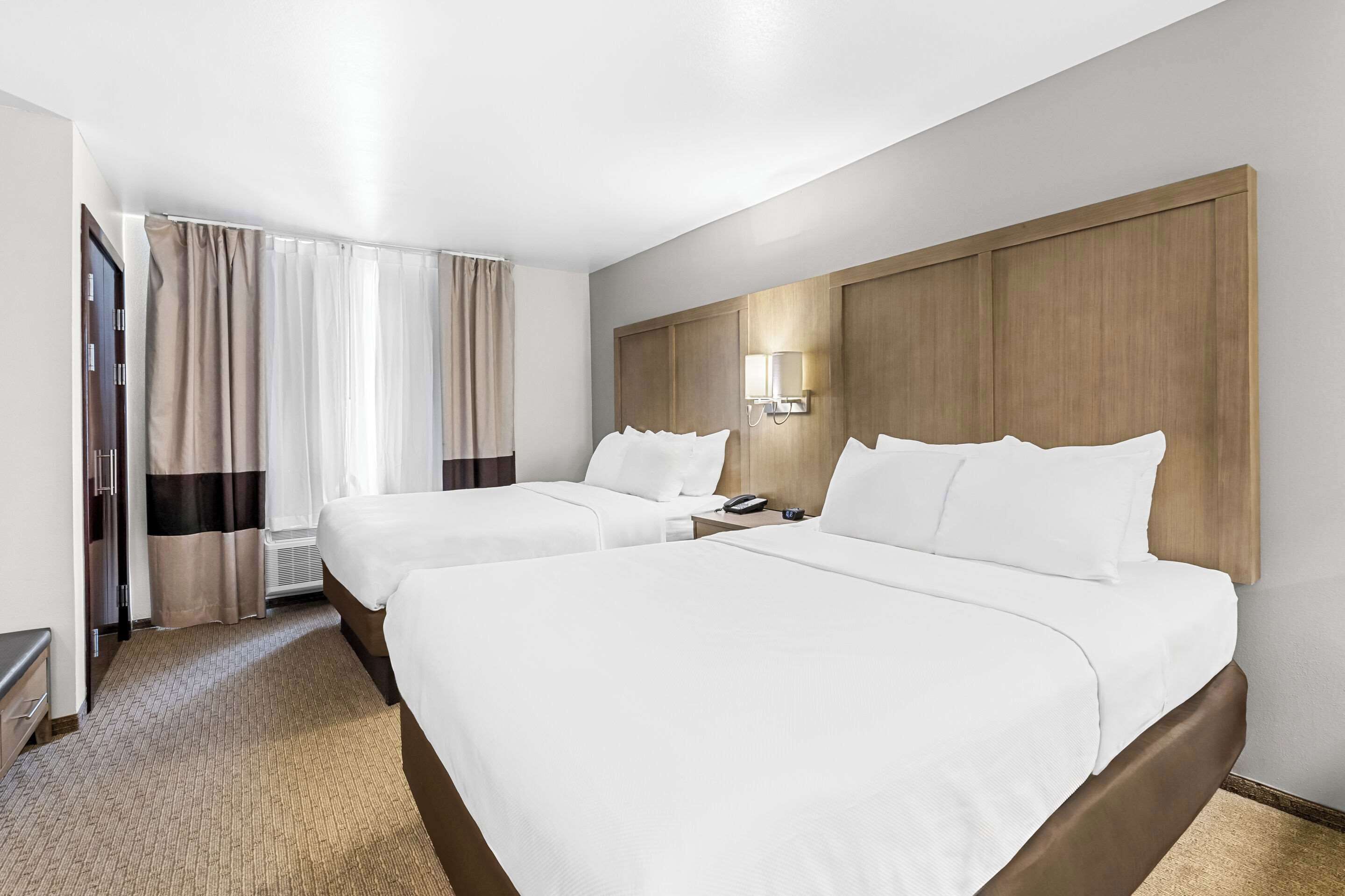 Quarto de hotel com duas camas, cabeceira de madeira, mesa de cabeceira com luminária e janela com cortinas.
