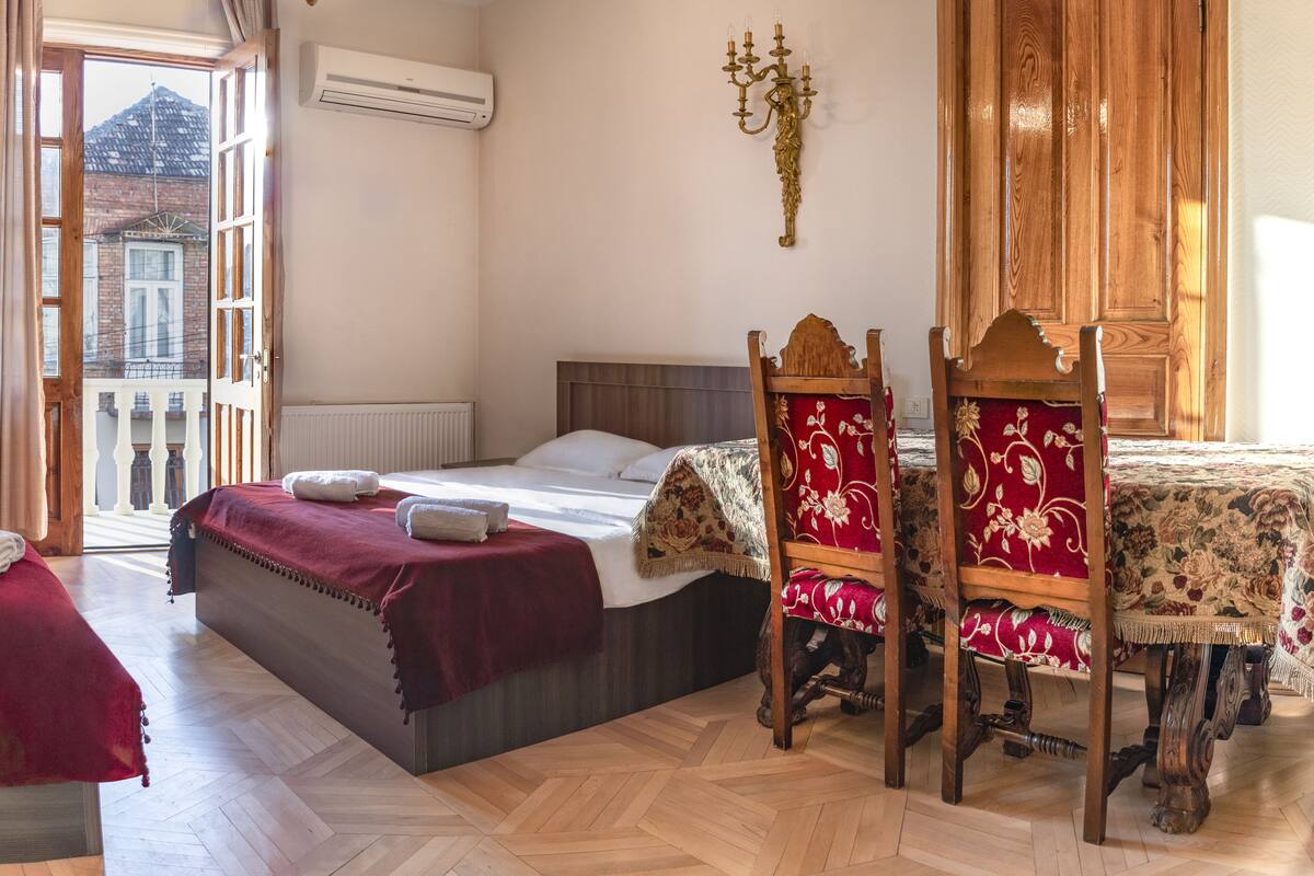 Ein Schlafzimmer mit einem Bett, einem Holzschrank, einer Kristallleuchtre und Blick auf ein Gebäude durch ein offenes Fenster.