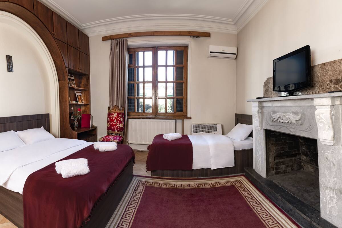 Ein Hotelzimmer mit zwei Betten, einem Kamin, einem Fernseher und einem großen Fenster mit Vorhängen.