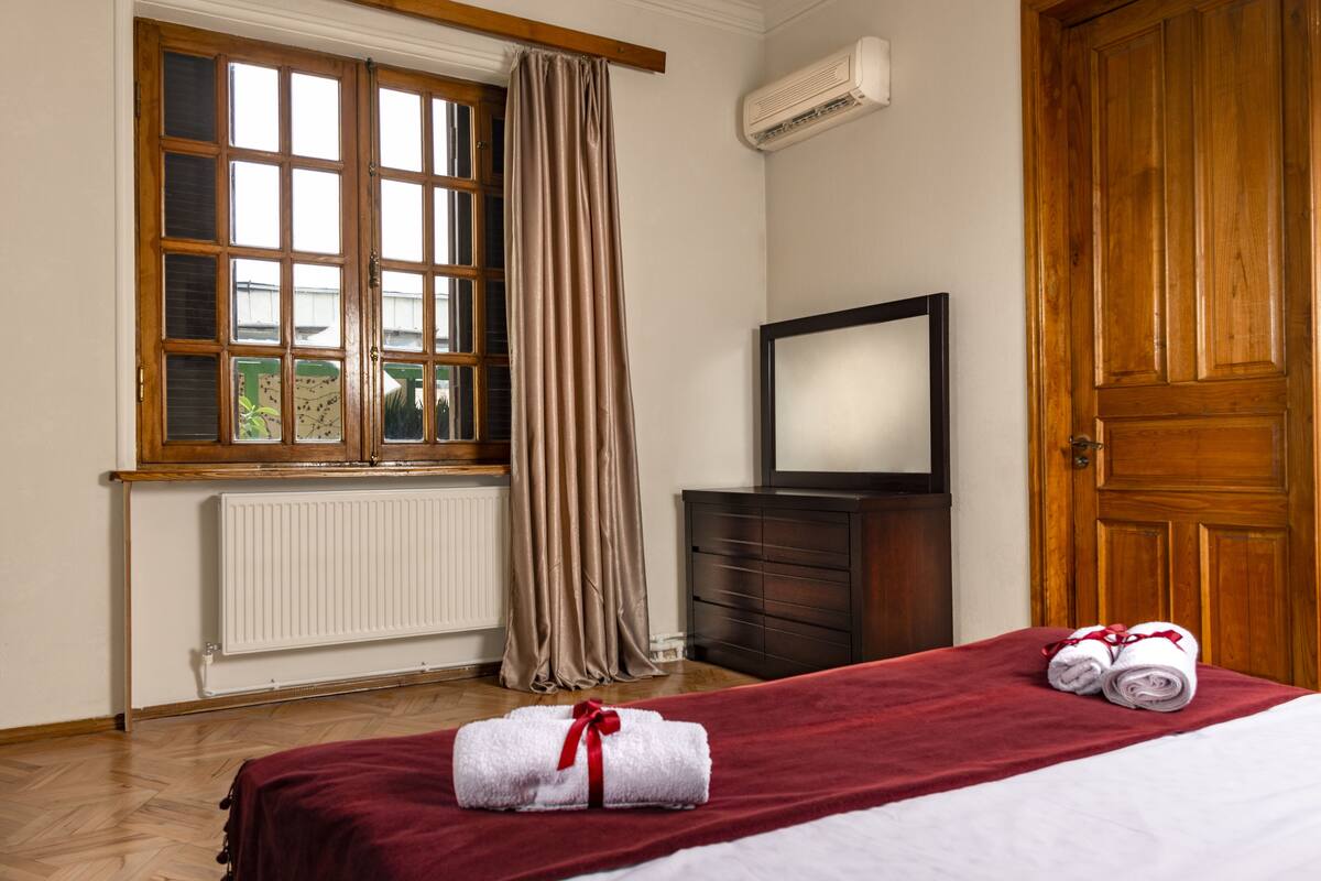 Ein Hotelzimmer mit Holzboden, einem großen Fenster mit Vorhängen, einem Heizkörper, einer Holzkommode, einem Spiegel und zwei aufgerollten Handtüchern auf dem Bett.