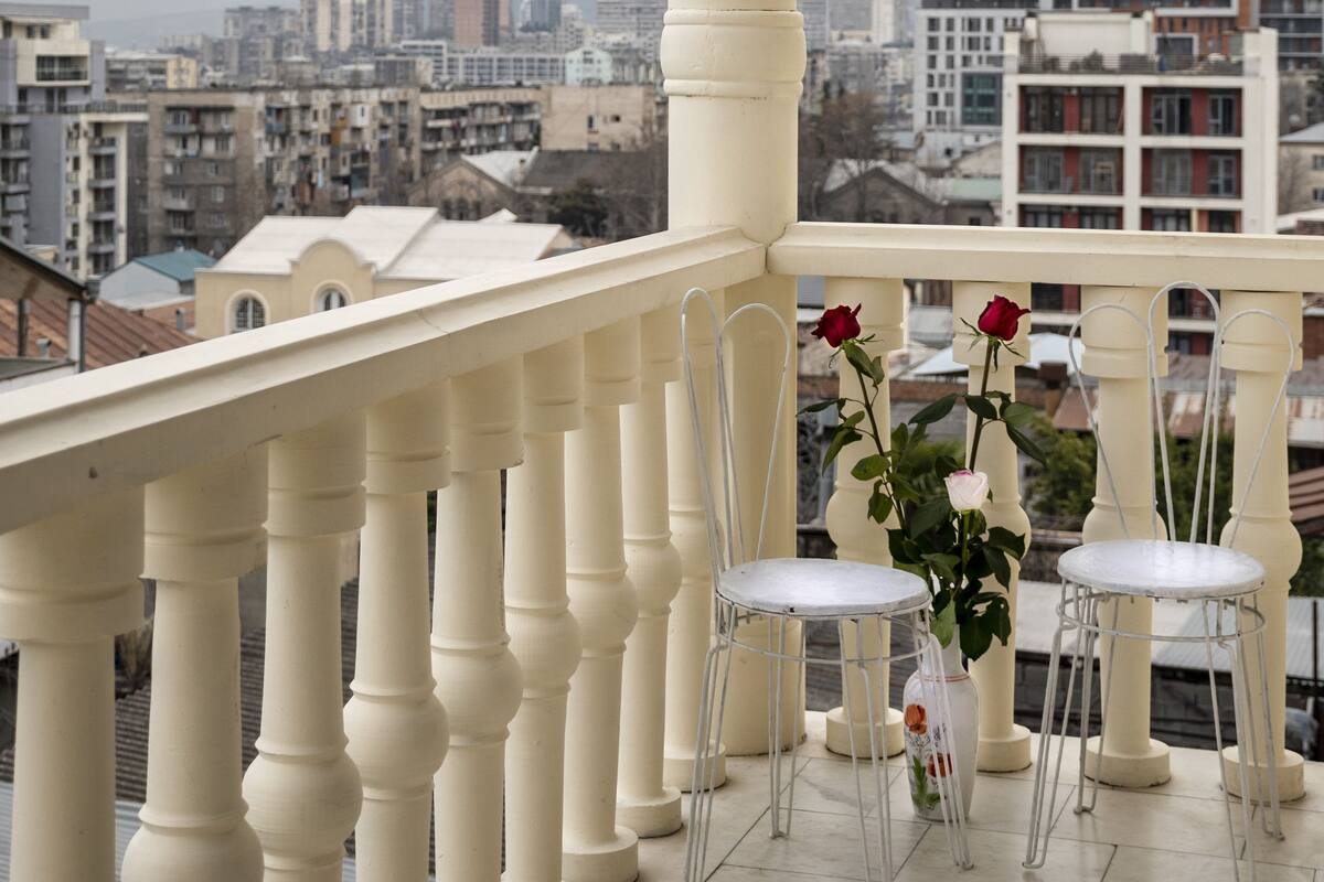 Ein Balkon mit weißen Geländern und Stühlen, der auf eine Stadtlandschaft mit Gebäuden und Bergen blickt.