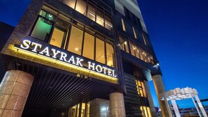 Exterior del hotel Stayrak con un llamativo letrero iluminado, amplias ventanas y un diseño arquitectónico moderno.