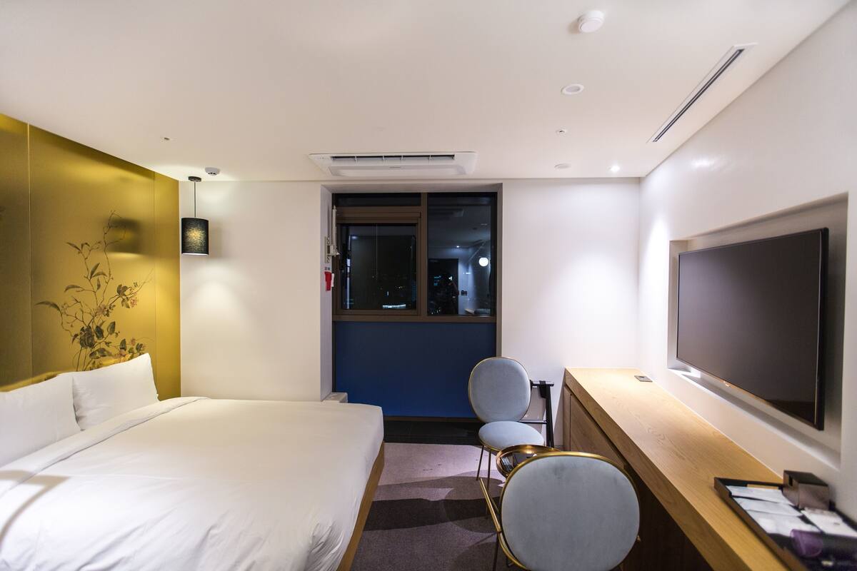 A hotel room with a bed, a desk with a flat-screen TV, a chair, and a window.